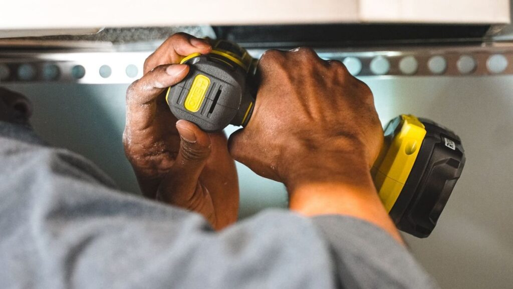 Mains tenant un tournevis électrique jaune noir sur une étagère métallique perforée
