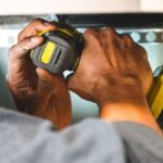 Mains tenant un tournevis électrique jaune noir sur une étagère métallique perforée