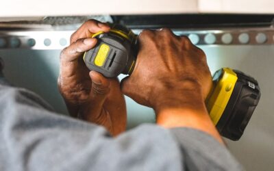 Mains tenant un tournevis électrique jaune noir sur une étagère métallique perforée