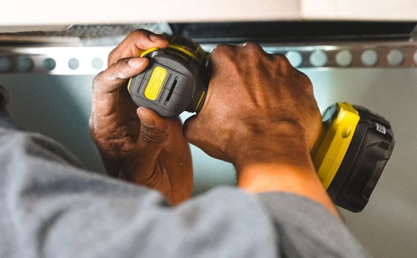 Mains tenant un tournevis électrique jaune noir sur une étagère métallique perforée
