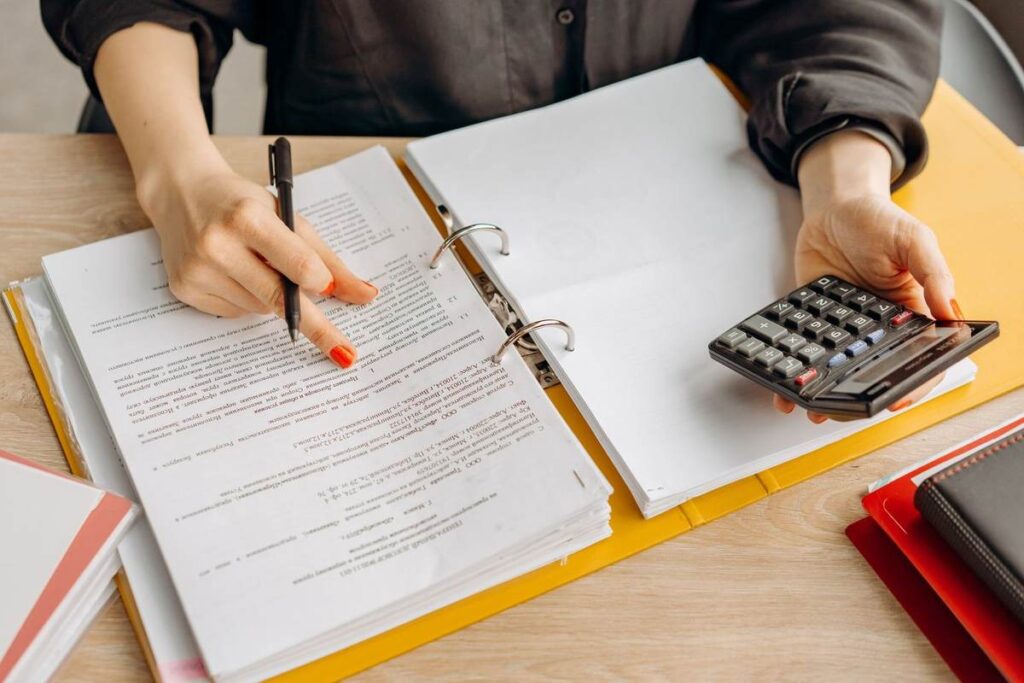 Une personne écrivant dans un classeur avec un stylo, tenant une calculatrice, sur une table